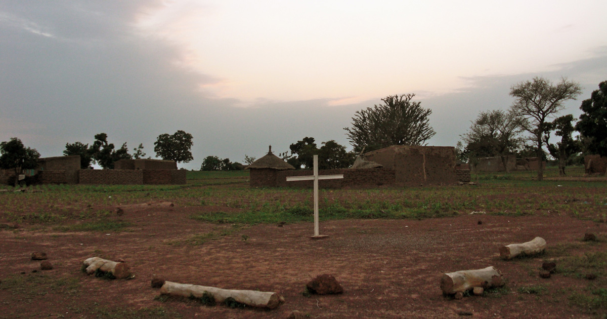 A simple cross sits in the ground with buildings in the background.