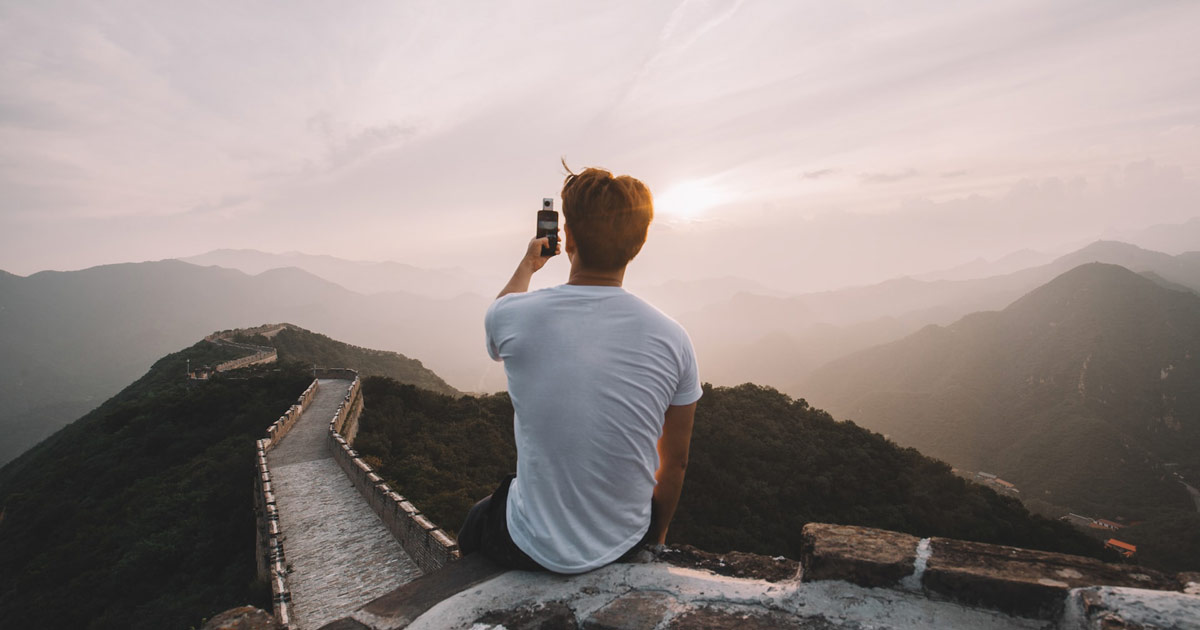 A young man sitting on the Great Wall of China, taking a picture with his cell phone.