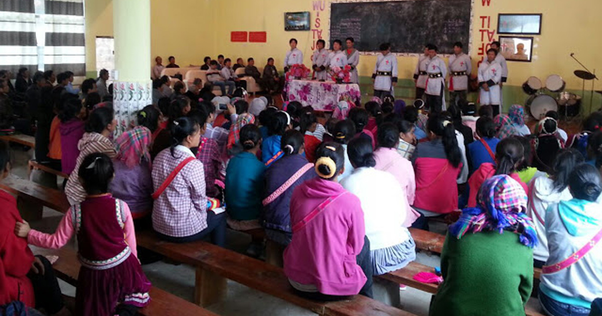 An audience is seated on wooden benches facing some individuals at the front of the meeting.