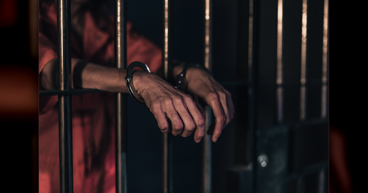 Man's hands extending from prison bars with handcuffs on his wrists.