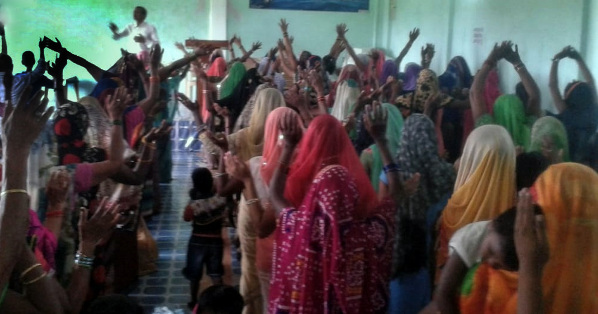 Worshippers are gathered in a small church. Many have their hands raised.