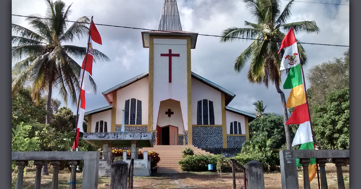 Trees surround a church building behind an open gate. There are colourful flags on either side of the gate.