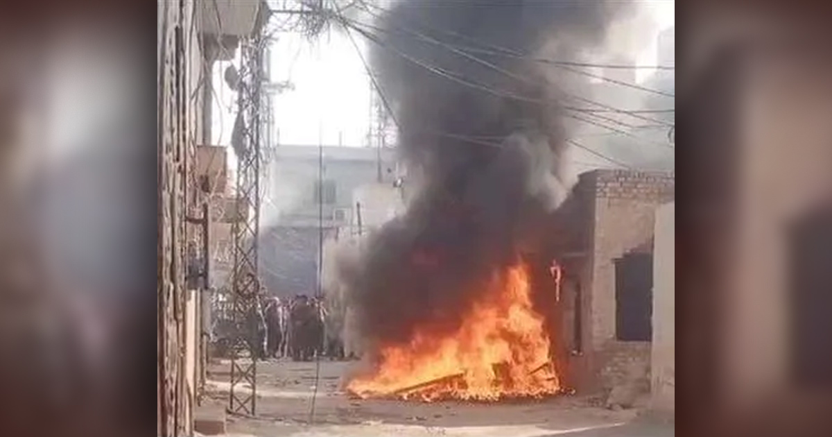 A fire is burning in the street. People are milling about in the background.