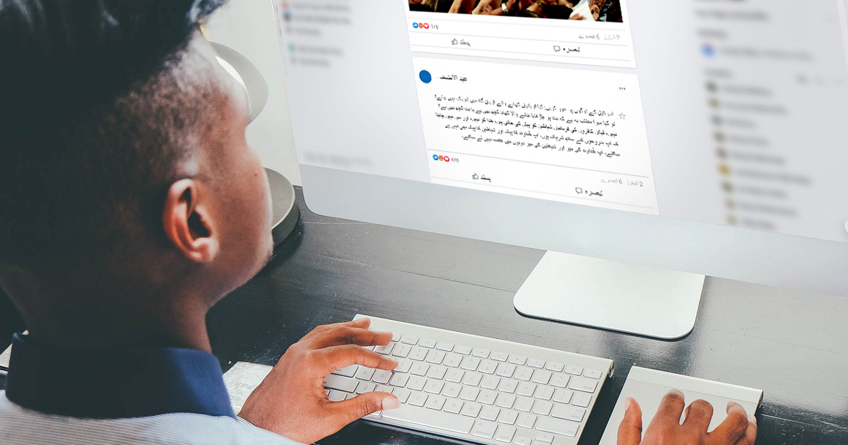 A man sitting at a desk, reading a Facebook post with 1 Corinthians 10: 18-21 in Urdu.