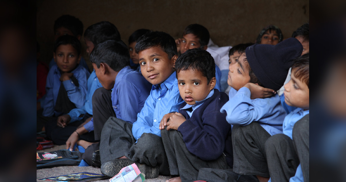 A group of schoolboys is sitting on the floor. Some are looking at the camera, others are looking at the teacher.