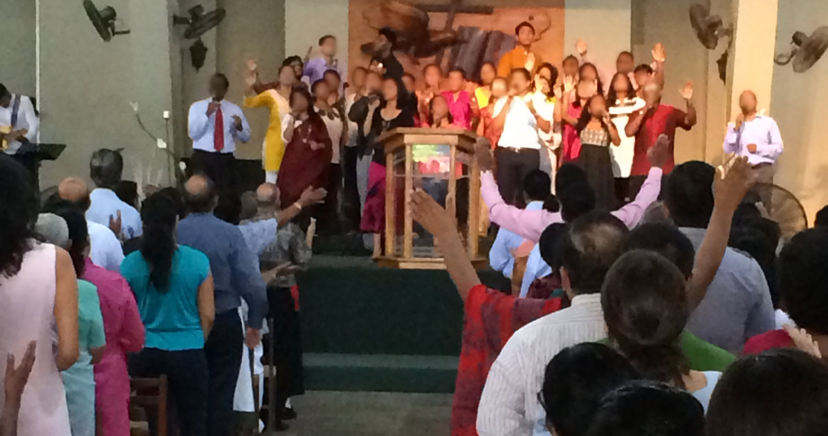 A congregation and a group at the front are worshipping the Lord together, many with their hands raised. 