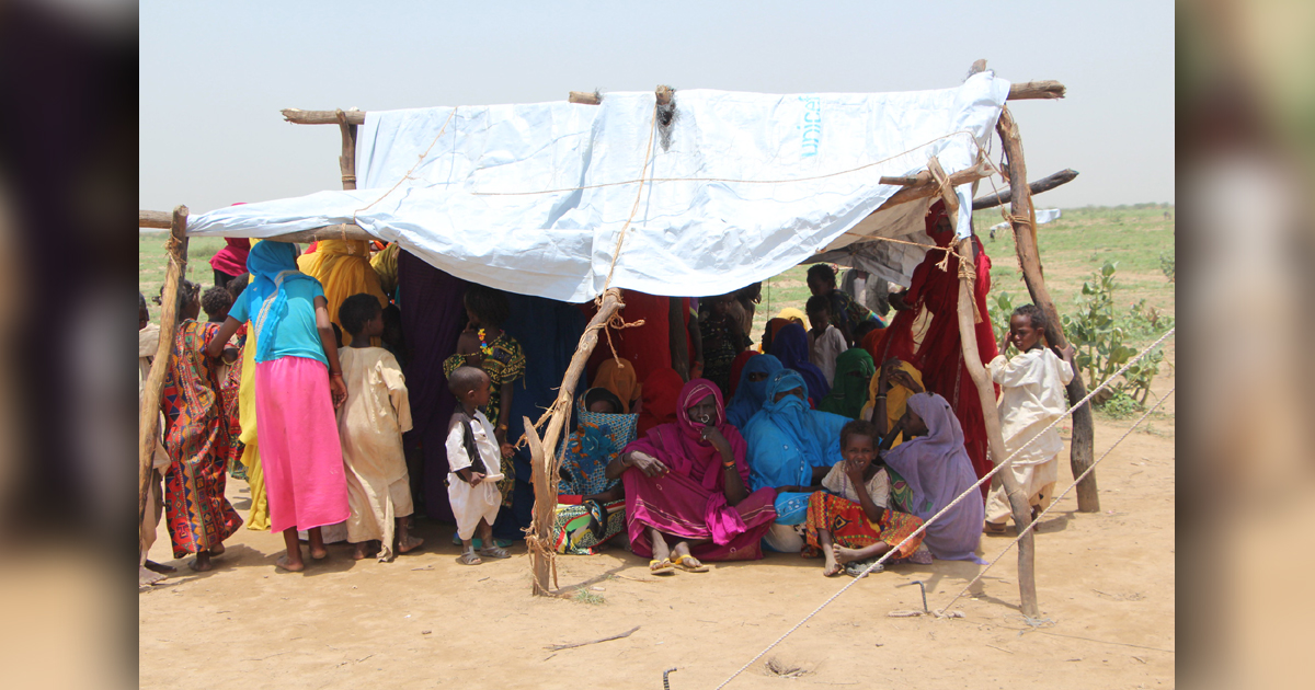 A group of people are gathered closely together under a rudimentary shelter made of poles and white cloth.