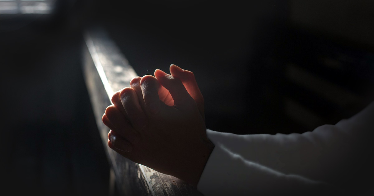 Praying hands resting against a rail. The sun from a window highlights the rail and hands in a dark room.